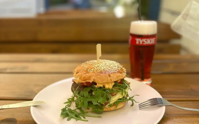 Burger z rukolą i sosem na białym talerzu z nożem i widelcem, obok szklanka piwa