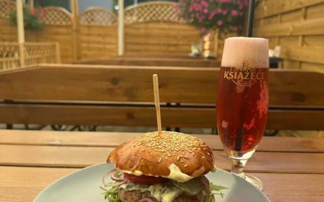 Burger z sezamową bułką, kotletem, serem pleśniowym i rukolą, obok szklanka piwa, w tle ogródek