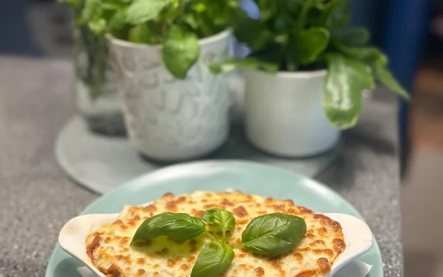Zapiekana lasagne z rozpuszczonym serem i listkami bazylii, w tle zioła i kwiaty w doniczkach