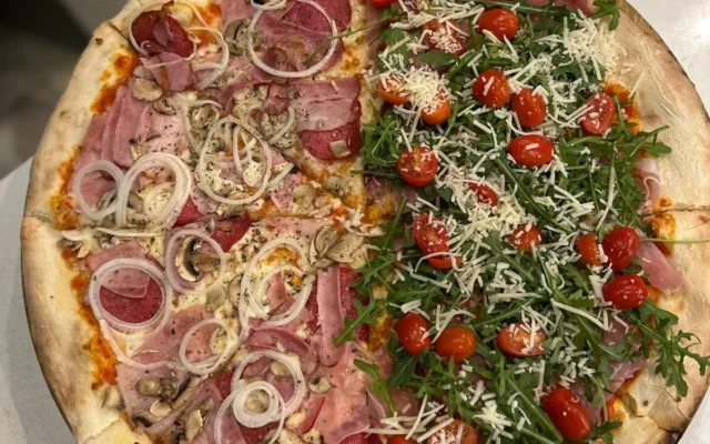 Pizza podzielona na pół: po lewej szynka, salami, pieczarki i cebula, po prawej rukola, pomidorki koktajlowe i parmezan na drewnianej desce